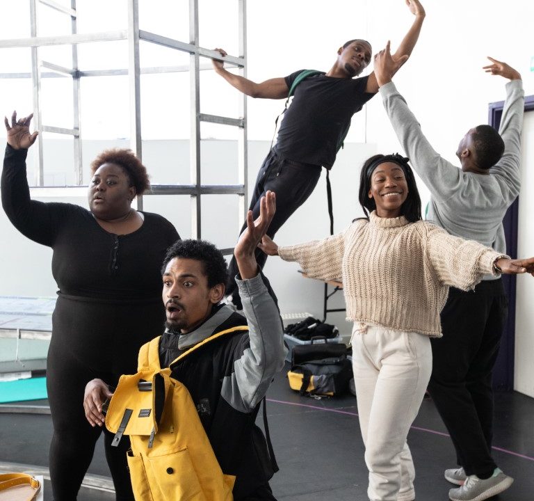 A man with a yellow backpack on the floor kneeling with his arm up. In the background is scaffolding and four people posing in ballet positions.