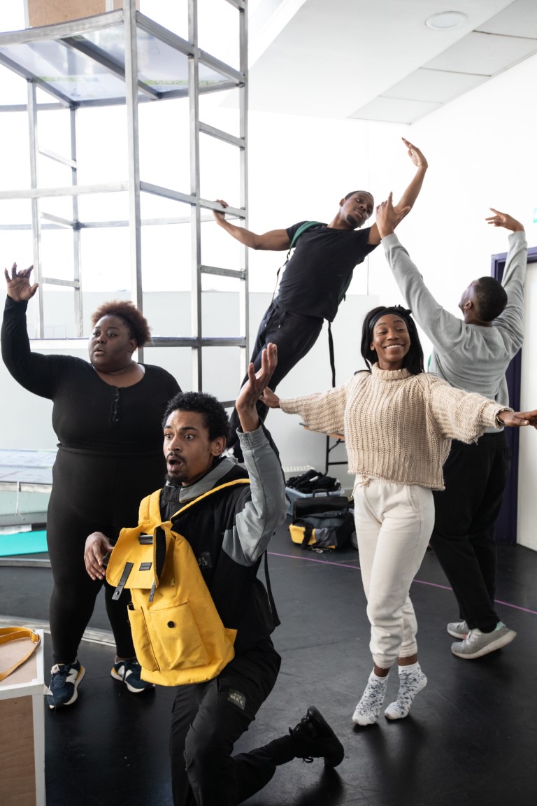 A man with a yellow backpack on the floor kneeling with his arm up. In the background is scaffolding and four people posing in ballet positions.