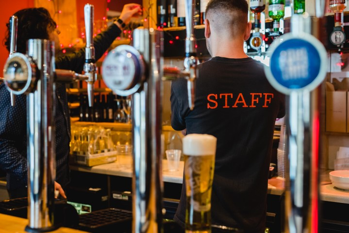 Someone working behind a bar wearing a T-shirt which says STAFF, with a pint of beer in the foreground.