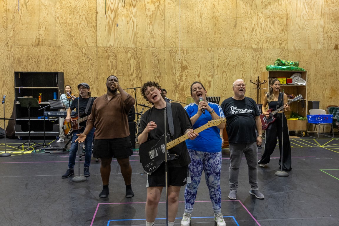 7 actors in rehearsals for Rapunzel, they are standing in a triangle formation singing into microphones, some are also playing instruments (keyboard and guitars)